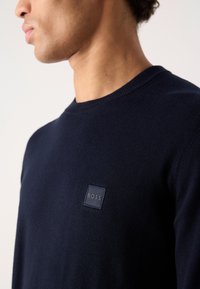 Navy blue knitted sweater with a round neckline and ribbed cuffs. Features a small square logo patch on the front. Soft texture.