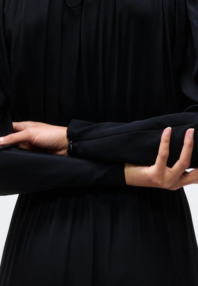 Χέρια σταυρωμένα στη μέση μπροστά από ένα μαύρο ένδυμα με μακριά μανίκια και λεπτομέρεια από πλισέ ύφασμα.