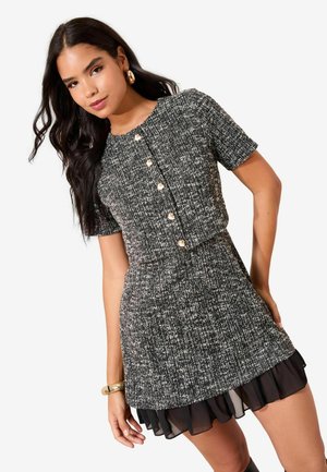 Melna un balta tvīda kleita ar īsām piedurknēm, apaļu izgriezumu, zelta pogām un kārtainu caurspīdīgu volānu apakšmalu.