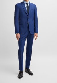 Niebieski garnitur o wąskim kroju, z klapami z wycięciami, dwoma guzikami i pojedynczą kieszenią na piersi. Połączony z jasnoniebieską koszulą i czarnymi butami.