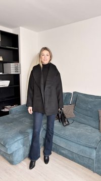 Melns pārmēru liels mētelis ar šāla apkakli, valkāts pār melnu augstu kaklu džemperi, kombinēts ar izplestām tumšām džinsa biksēm un melniem potīšu zābakiem.