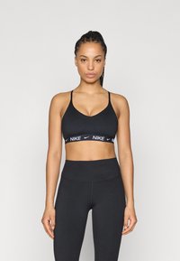 Melnā sporta krūšturis ar regulējamām lencēm un platu zīmolu joslu ar baltu uzrakstu. Kopā ar atbilstošām augsta jostasvietas melnām legingām.