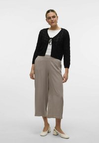 Cardigan nero a maglia con una cintura in vita, abbinato a pantaloni beige a gamba larga. Il modello indossa una maglietta bianca e scarpe di colore chiaro.