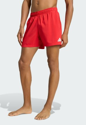 Uomo che indossa pantaloncini sportivi rosso brillante con logo Adidas bianco sulla gamba sinistra, in piedi a piedi nudi su uno sfondo semplice.