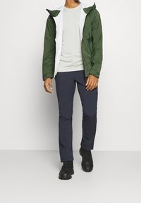 Zielona i biała wodoodporna kurtka, szara koszula, granatowe spodnie i czarne buty. Kurtka ma kaptur i zapinane na suwak mankiety.