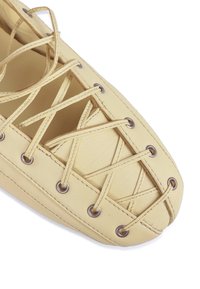 Sárga bőrcipő hegyes orral, keresztezett fűződizájnnal és fém szemekkel a fűzőhöz. Simább tapintás, minimál stílus.