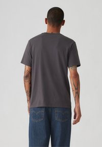 Tricou gri din bumbac cu mâneci scurte, decolteu rotund și o croială lejeră. Prezintă tatuaje vizibile pe brațe și blugi largi de culoare albastră.