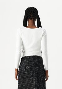 Haut blanc à manches longues en maille côtelée avec un décolleté en trompette, associé à une jupe à carreaux noir et blanc. Les matériaux sont en coton et en tissu texturé.