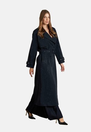 Frau in einem langen, taillierten schwarzen Coat mit Nadelstreifen und passenden, weit geschnittenen Hosen sowie schwarzen hohen Schuhen, die vor einem schlichten Hintergrund läuft.