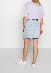 Jupe en denim bleu clair avec un ourlet brut, cinq poches et un patch logo sur la ceinture arrière, associée à un T-shirt violet pastel et des baskets blanches.