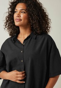 Camicia nera a bottoni con vestibilità rilassata, maniche corte e una consistenza morbida. Presenta un colletto a punte e dettagli di bottoni discreti.
