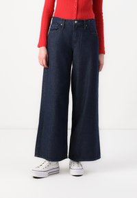 Jean en denim foncé à jambe large et taille haute, avec des poches avant et des coutures ton sur ton. Associé à des baskets blanches. Haut en maille rouge visible.