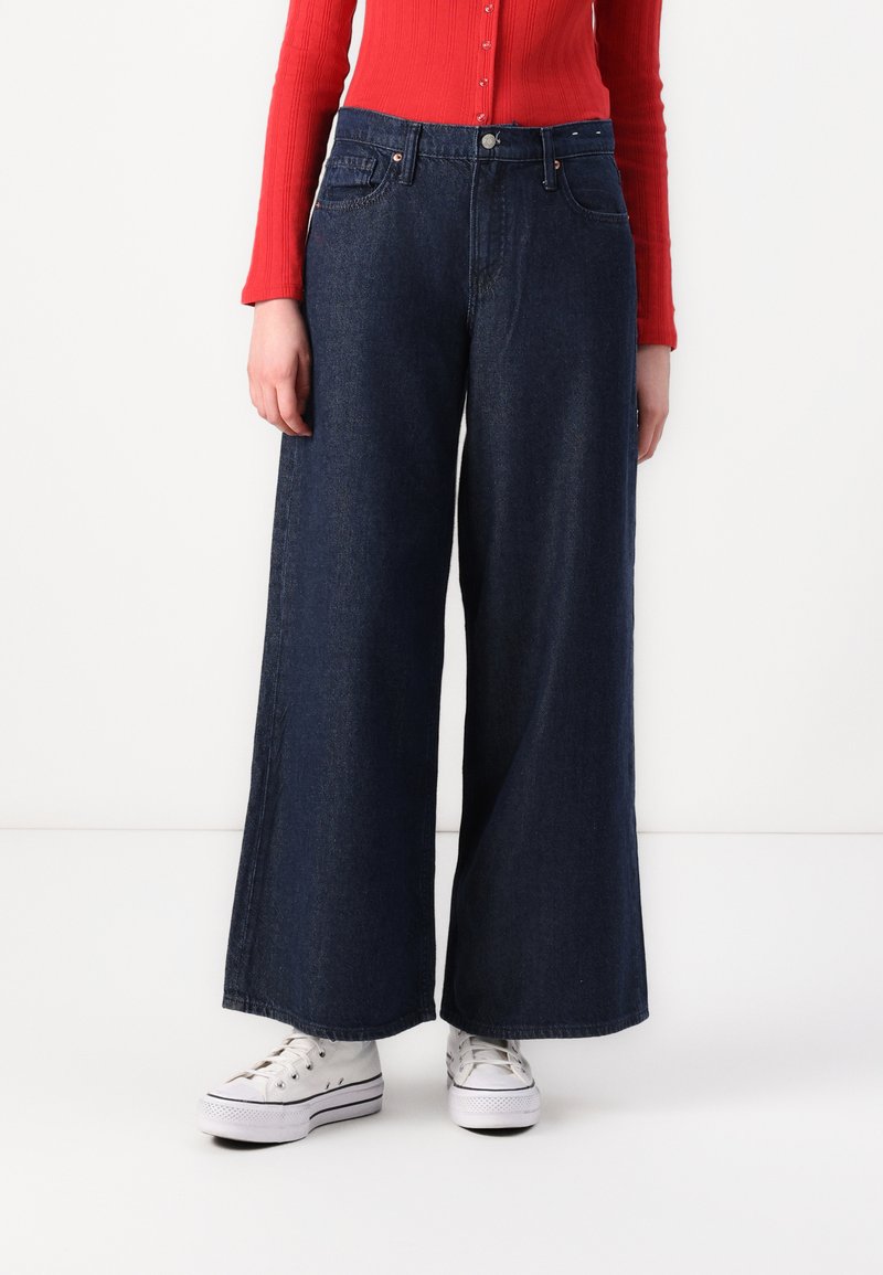 Jean en denim foncé à jambe large et taille haute, avec des poches avant et des coutures ton sur ton. Associé à des baskets blanches. Haut en maille rouge visible.