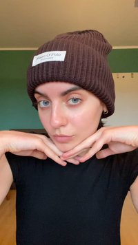 Strikket brun hue med et hvidt label, der har en ribbet tekstur; båret af en person i en sort skjorte med hænderne placeret under hagen.