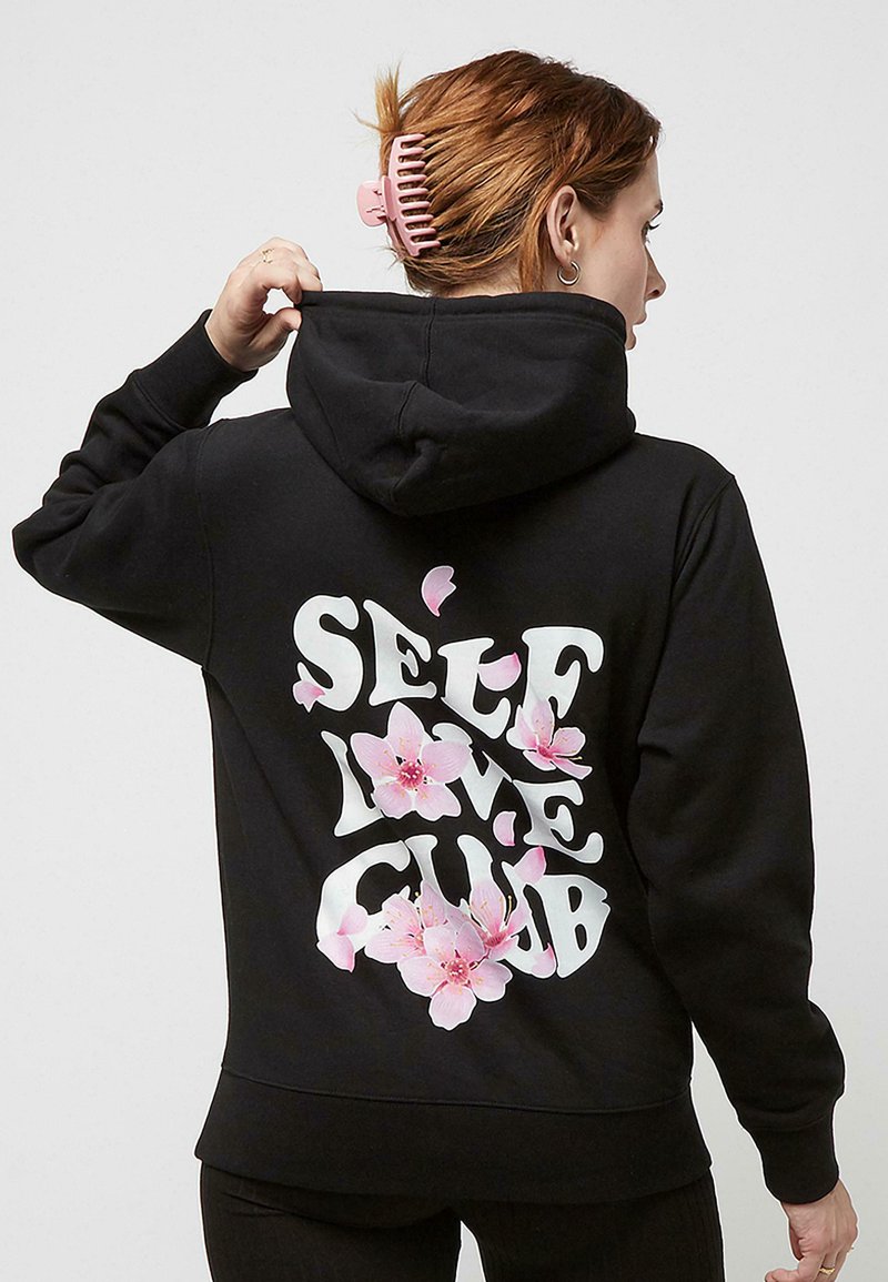 Hoodie noir avec un design textuel blanc "SELF LOVE CLUB" agrémenté d'accents floraux roses, mettant en avant un mélange de coton et une coupe décontractée.