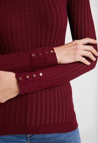Burgundy ribbed neulepaita, jossa on tiukat hihat, joissa jokaisessa on kolme pronssista nappia, ja tiukka vyötärösuunnittelu.