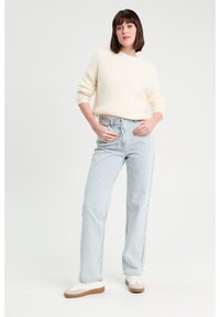 Pull en tricot de couleur crème avec des manches longues, associé à un jean taille haute à rayures bleu clair et des baskets blanches à semelles beige.
