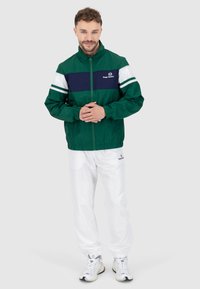 Grün und marinefarbener, farblich abgesetzter Windbreaker mit weißen Streifen. Leichter Stoff, Reißverschluss vorne, elastische Bündchen. Getragen mit weißen Hosen und Sneakers.