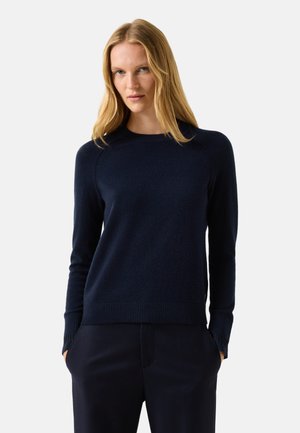 Kvinde med langt blondt hår iført en tætsiddende marineblå langærmet sweater og matchende marineblå bukser med hænderne i lommerne.