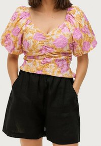 Blumenbluse mit rosa und gelben Rosen auf weißem Hintergrund, mit Puffärmeln und tailliertem Schnitt, kombiniert mit schwarzen, hochgeschnittenen Shorts.