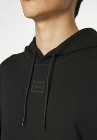 Hoodie noir en tissu doux avec une capuche à cordon de serrage, présentant un patch logo texturé sur la poitrine et une finition lisse.