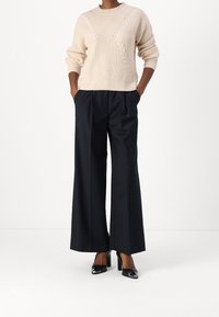 Pull en tricot beige avec un motif de câble texturé, associé à un pantalon large bleu marine et des chaussures à talons hauts noires.