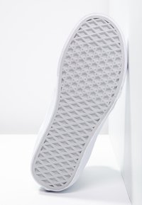 Semelle d'une basket blanche avec un motif en losanges, positionnée verticalement contre un coin blanc entre un mur et le sol.