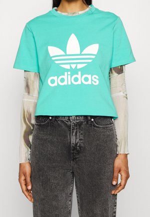 Turkis bomulds-crop t-shirt med hvid Adidas-logo og trefoil-design. Båret over en langærmet mønstret bluse og parret med sorte jeans.