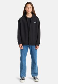Ragazzina in piedi, che indossa una felpa nera con zip, jeans blu e sneakers bianco-nere, su uno sfondo chiaro e neutro.