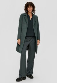 Dunkelgrüner Wollmantel mit Kragen, Ein-Knopf-Verschluss und zwei seitlichen Taschen, getragen über einem schwarzen Oberteil und grauen Hosen. Dicke schwarze Plateauschuhe.