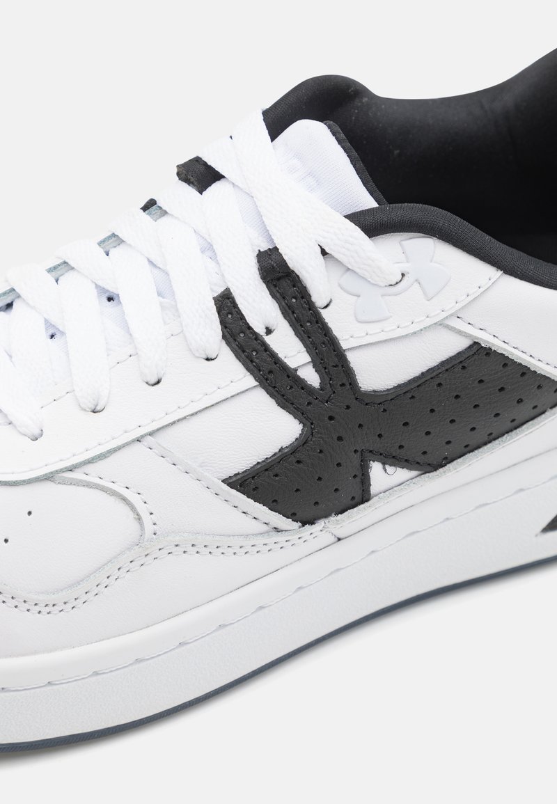 Zapatilla de cuero blanca con acentos perforados en negro, cordones planos blancos y suela de goma texturizada. Presenta un logo en el costado.