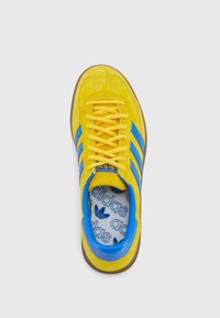 Zapatilla de ante amarilla con diseño de tres rayas azules, cordones amarillos y acento en el talón azul. Plantilla blanca con patrón de logo de Adidas en azul.