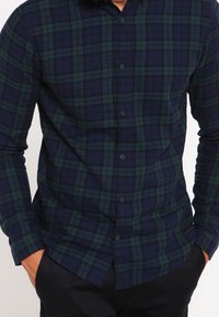 Marineblauw en groen geruit flanellen shirt met lange mouwen, knoopsluiting aan de voorkant, zachte textuur en een slanke pasvorm, gepresenteerd met handen in de zakken.