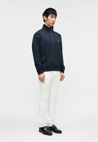 Σκούρο μπλε φούτερ με φερμουάρ στον λαιμό, ριμπ μανσέτες και μπροστινή τσέπη, συνδυασμένο με λευκό παντελόνι με στενή μπότα και μαύρα loafer.