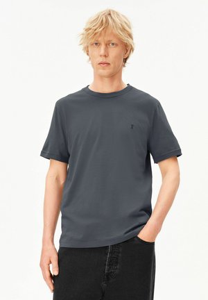 Blonder Mann trägt ein schlichtes dunkelgraues T-Shirt mit kurzen Ärmeln und schwarze Jeans, steht mit einer Hand in der Tasche vor weißem Hintergrund.
