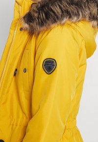 Parka amarilla con capucha forrada de piel sintética, que cuenta con dos bolsillos frontales y una insignia en la manga, fabricada con tejido resistente al agua.