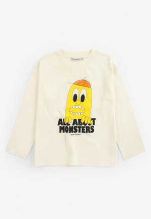 Kremowa koszulka z długim rękawem z żółtym, kreskówkowym potworem w pomarańczowej czapce i czarnym napisem ""All About Monsters"" na przodzie.