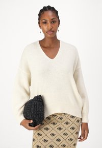 Kremowy, oversize'owy sweter z dekoltem w serek, wykonany z miękkiego materiału, w połączeniu z fakturowanym czarnym kopertówką oraz wzorzystą beżową spódnicą z czarnymi akcentami.