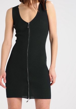 Abito nero senza maniche in maglia a coste con cerniera frontale, silhouette aderente e scollo a V indossato da una donna su uno sfondo semplice.