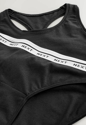 Fekete sportmelltartó, hátul versenypályára tervezett kialakítással, puha pamut anyagból, és egy fehér pánttal, amelyen az "NEXT" szó ismétlődik az elején.