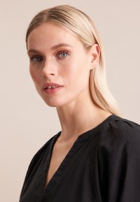 Blusa nera a scollo a V con maniche corte a sbuffo, realizzata in tessuto testurizzato. Il modello presenta un trucco minimal e capelli biondi lisci e lucenti.