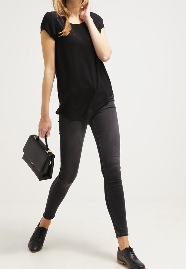 Femme portant un haut noir, un jean skinny foncé, des chaussures noires, tenant un sac à main noir, posant devant un fond uni.