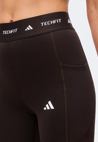 Κοντινό πλάνο μαύρου leggings Adidas Techfit με λευκό λογότυπο και ψηλό ζωνάκι με κείμενο "Techfit" και σύμβολα Adidas.