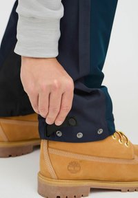 Waterproof navy pants being adjusted at the ankle, featuring snap closures; worn with brown work boots with yellow laces and white stitching.