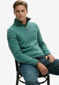 Turkusowy sweter z zamkiem przy szyi, ze ściągaczami na mankietach, teksturowanym splotem i kratkowaną podszewką przy kołnierzu, zestawiony z niebieskimi dżinsami. Modelka siedzi na czarnym krześle.