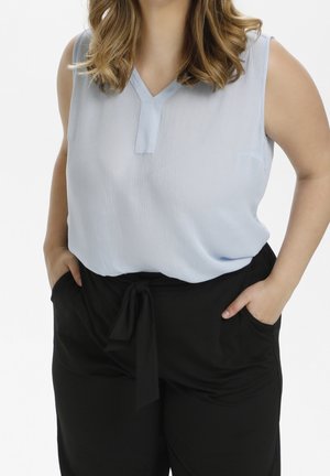 Kobieta w bezrękawowej jasnoniebieskiej bluzce wpuszczonej w wysokowalne czarne spodnie z wiązanym paskiem, z rękami w kieszeniach.