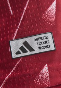 Λευκή και μαύρη ετικέτα Adidas "Authentic Licensed Product" σε υφασμάτινη κόκκινη επιφάνεια με λευκά διαγώνια σχέδια.