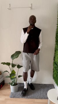 Man staat met een microfoon, draagt een bruine mouwloze shirt over een wit lange mouwen shirt, grijze korte broek en zwarte schoenen. Groene plant in de buurt.