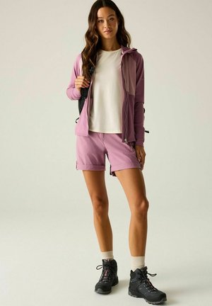 Frau trägt eine pinke Jacke und Shorts, weißes Hemd, beige Socken, schwarze Wanderschuhe und trägt einen schwarzen Rucksack, steht vor einem einfarbigen Hintergrund.