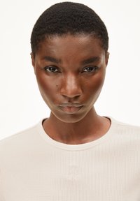 Lichtbeige lange mouw top van een gestructureerde stof, met een ronde halslijn en subtiele geborduurde logodetails aan de voorkant.